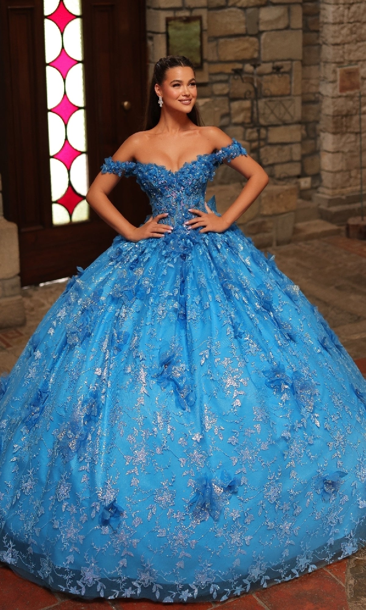 A woman poses indoors, smiling with her hands on her hips in the Amarra 54243 Off-Shoulder Quinceanera Dress, featuring lace details and a full skirt.
